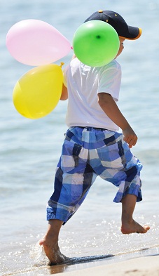 small boy wearing shorts running down the beach carrying three balloons