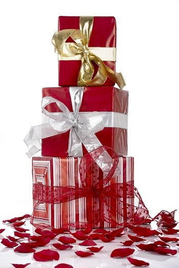 tack of christmas gifts wrapped in red and gold foil paper with large bows sitting in a pile of rose pedals